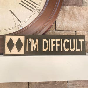May include: A rectangular wooden sign with the words "I'M DIFFICULT" carved in a light color. The sign also features a diamond-shaped ski slope symbol. The background is a dark brown color.
