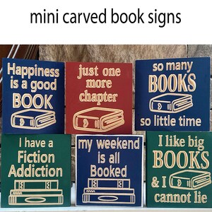 May include: Six wooden signs with book-themed sayings. The signs are painted in various colors, including blue, green, red, and gold. The signs feature sayings such as "Happiness is a good book", "Just one more chapter", "So many books, so little time", "I have a fiction addiction", "My weekend is all booked", and "I like big books & I cannot lie".