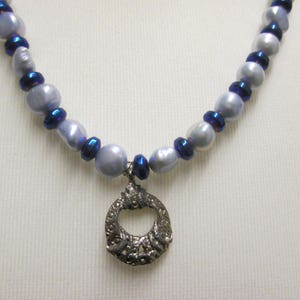 以下が含まれることがあります： 銀色のハート型のペンダントが付いた銀色のネックレス。ネックレスは白と青の虹色のビーズでできています。