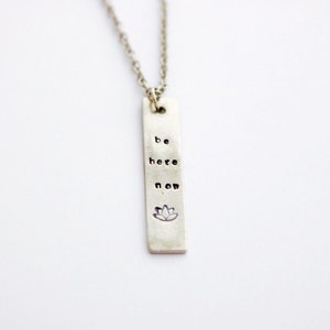 Op de afbeelding: Zilveren ketting met een rechthoekige hanger waarop "be here now" staat en een lotusbloem symbool.
