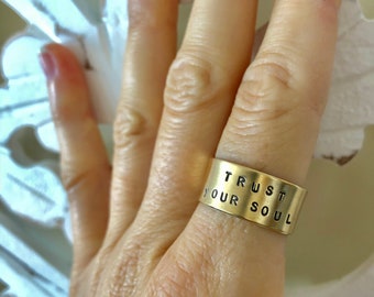 Puede incluir: Un anillo dorado con las palabras "TRUST YOUR SOUL" estampadas en el exterior. El anillo es ancho y envuelve un dedo. El fondo es blanco y blanquecino borroso.