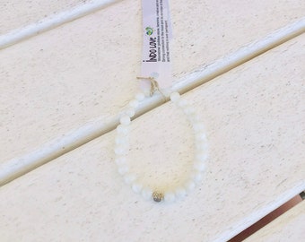 May include: White moonstone beaded bracelet with a silver charm. The bracelet is on a white wooden surface. The text on the card reads "INDO-LOVE Moonstone goddess stone feminine, create and encon Strong connection to the moon and its minder of the cycle and that nothing is ever permanent."