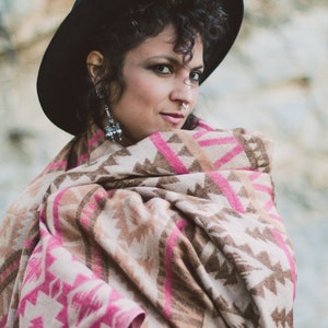 May include: A woman wearing a black hat and a patterned shawl. The shawl features a geometric design in shades of pink, brown, and beige. The shawl is wrapped around the shoulders.