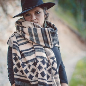 May include: A person wearing a black fedora hat and a beige and black patterned scarf. The scarf has a geometric design and is draped around the neck. The person is also wearing bracelets and a black long-sleeve shirt.