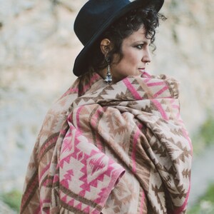 May include: A woman wearing a black hat and a fringed shawl. The shawl features a geometric pattern in shades of pink, brown, and beige. The woman is wearing silver earrings.