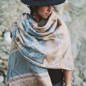 May include: A person wearing a black hat and a fringed shawl. The shawl features a geometric pattern in shades of blue, tan, and white. The person is also wearing a bracelet.