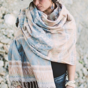 May include: A woman wearing a black hat and a patterned shawl in shades of blue, beige, and white. The shawl has a fringed edge. The woman is also wearing bracelets and earrings. The background is a light gray.
