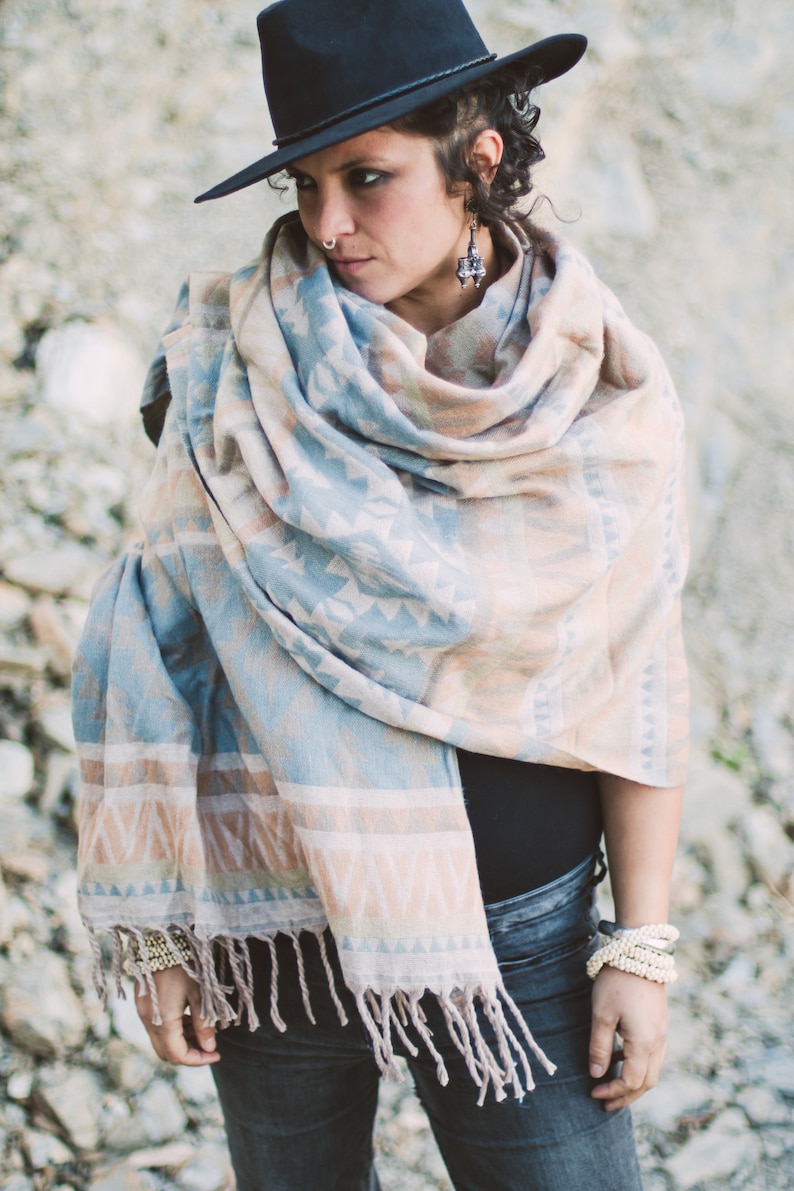 May include: A woman wearing a black hat and a patterned shawl in shades of blue, beige, and white. The shawl has a fringed edge. The woman is also wearing bracelets and earrings. The background is a light gray.