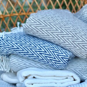 May include: A stack of three blankets in a wicker basket. The top blanket is gray with a white chevron pattern. The middle blanket is blue with a white chevron pattern. The bottom blanket is white with a blue stripe pattern.