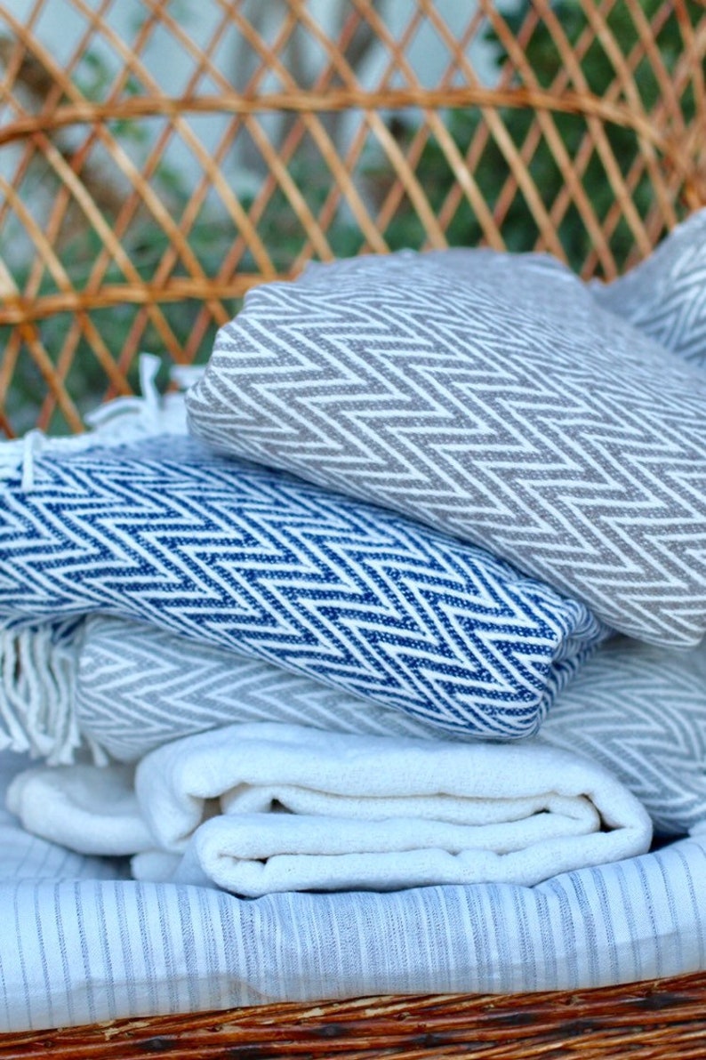May include: A stack of three blankets in a wicker basket. The top blanket is gray with a white chevron pattern. The middle blanket is blue with a white chevron pattern. The bottom blanket is white with a blue stripe pattern.