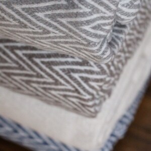 May include: A stack of three blankets with different geometric patterns. The top blanket is gray and white with a chevron pattern, the middle blanket is brown and white with a zig-zag pattern, and the bottom blanket is white with a blue stripe.