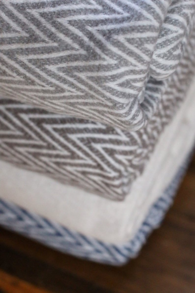 May include: A stack of three blankets with different geometric patterns. The top blanket is gray and white with a chevron pattern, the middle blanket is brown and white with a zig-zag pattern, and the bottom blanket is white with a blue stripe.