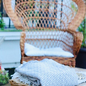 May include: A gray and white striped throw blanket folded on top of a wicker ottoman. The ottoman is in front of a wicker peacock chair.