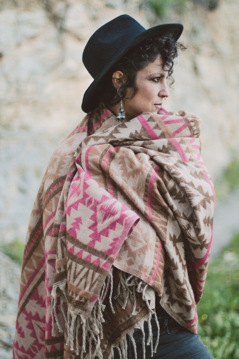May include: A woman wearing a black hat and a fringed shawl. The shawl features a geometric pattern in shades of pink, brown, and beige. The woman is wearing silver earrings.