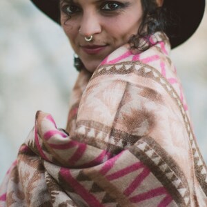 May include: A woman wearing a black hat and a patterned shawl. The shawl features a geometric design in shades of pink, brown, and beige. The woman has dark curly hair and a nose ring.