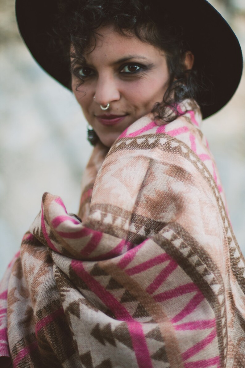 May include: A woman wearing a black hat and a patterned shawl. The shawl features a geometric design in shades of pink, brown, and beige. The woman has dark curly hair and a nose ring.