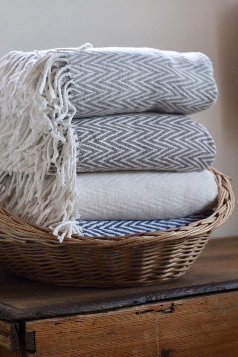 May include: A wicker basket filled with four folded blankets. The blankets are in a chevron pattern and are in shades of gray, white, and blue.