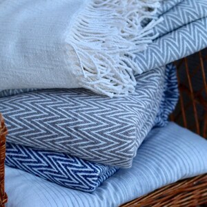 May include: A stack of three blankets with different patterns. The top blanket is white with a fringe, the middle blanket is gray with a chevron pattern, and the bottom blanket is blue with a chevron pattern. The blankets are sitting on a wicker chair.