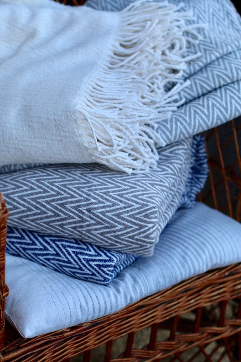 May include: A stack of three blankets with different patterns. The top blanket is white with a fringe, the middle blanket is gray with a chevron pattern, and the bottom blanket is blue with a chevron pattern. The blankets are sitting on a wicker chair.