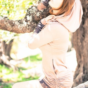 May include: A woman wearing a light brown hooded sweatshirt with a drawstring at the waist. The sweatshirt has a lace trim at the bottom. She is standing in a forest with trees in the background.