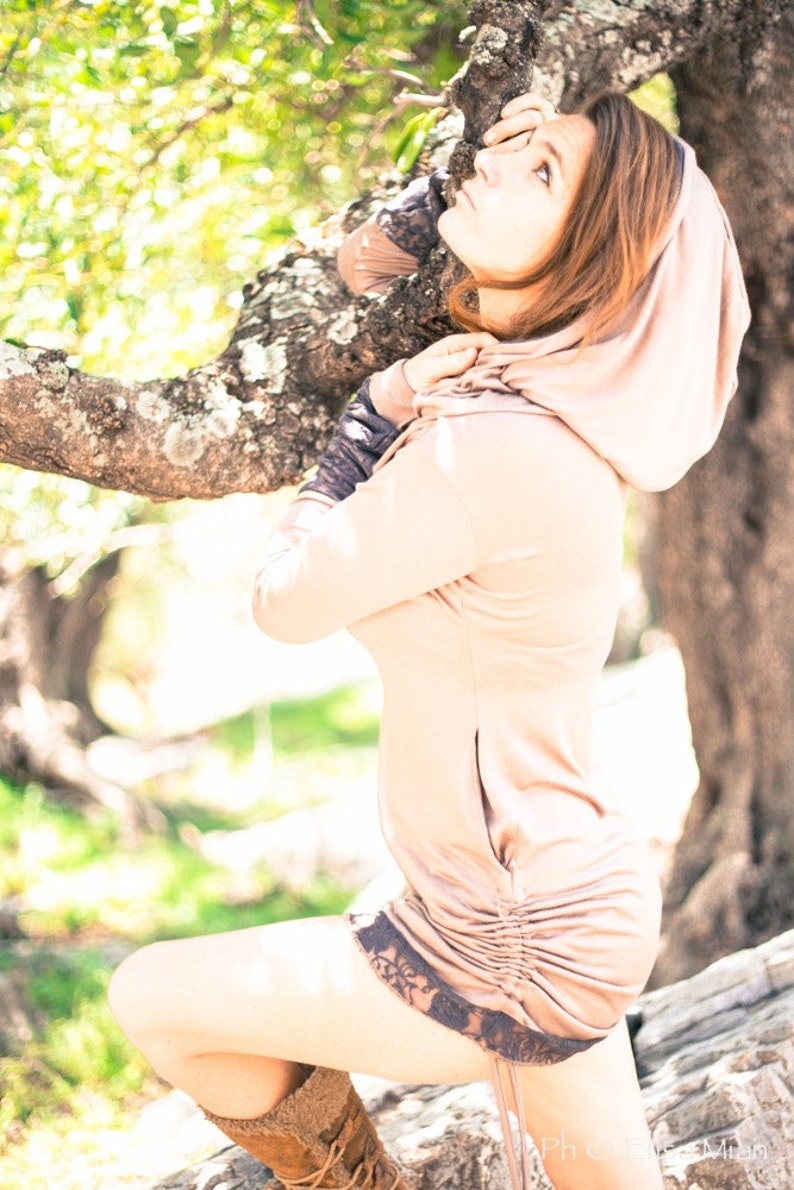 May include: A woman wearing a light brown hooded sweatshirt with a drawstring at the waist. The sweatshirt has a lace trim at the bottom. She is standing in a forest with trees in the background.