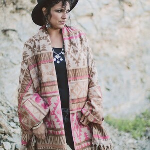 May include: A woman wearing a black hat and a patterned shawl with pink and brown geometric designs. The shawl has a fringed hem. She is also wearing a black top, silver jewelry, and bracelets.