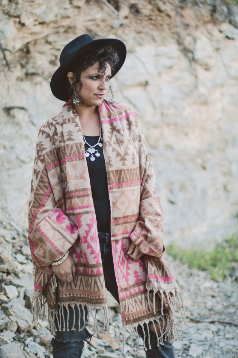 May include: A woman wearing a black hat and a patterned shawl with pink and brown geometric designs. The shawl has a fringed hem. She is also wearing a black top, silver jewelry, and bracelets.