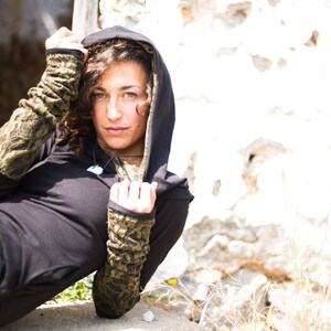 May include: A woman wearing a black hooded top with green and brown patterned sleeves. The hood is pulled up over her head and she is looking at the camera.