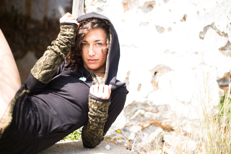 May include: A woman wearing a black hooded top with green and brown patterned sleeves. The hood is pulled up over her head and she is looking at the camera.
