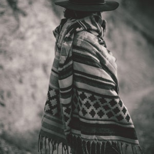 May include: A person wearing a black hat and a patterned poncho with fringe. The poncho features a geometric design in shades of gray and white. The image is in black and white.