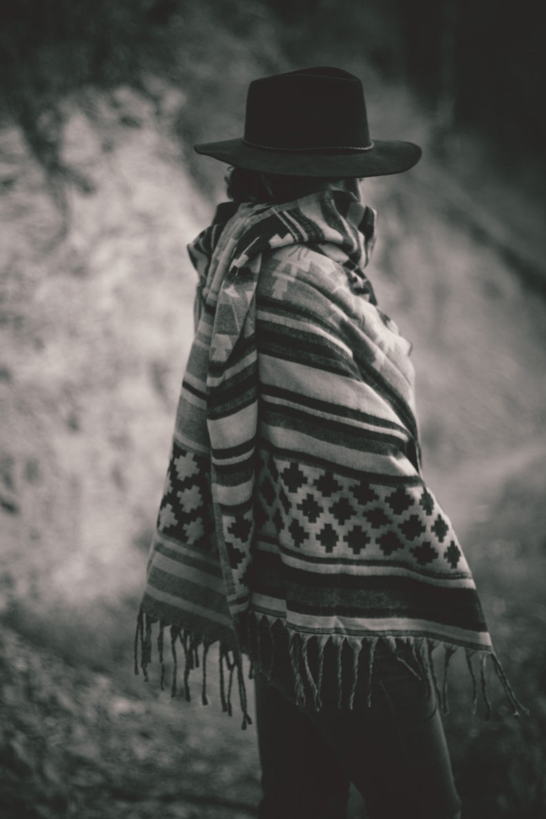 May include: A person wearing a black hat and a patterned poncho with fringe. The poncho features a geometric design in shades of gray and white. The image is in black and white.