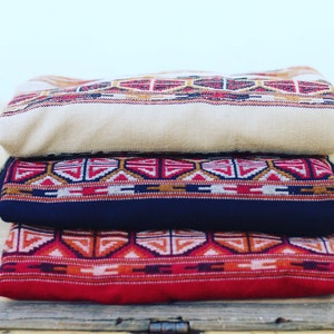 May include: Three woven blankets stacked on top of each other. The top blanket is beige with a red and brown geometric pattern. The middle blanket is dark blue with a red, white, and brown geometric pattern. The bottom blanket is red with a white, orange, and brown geometric pattern.