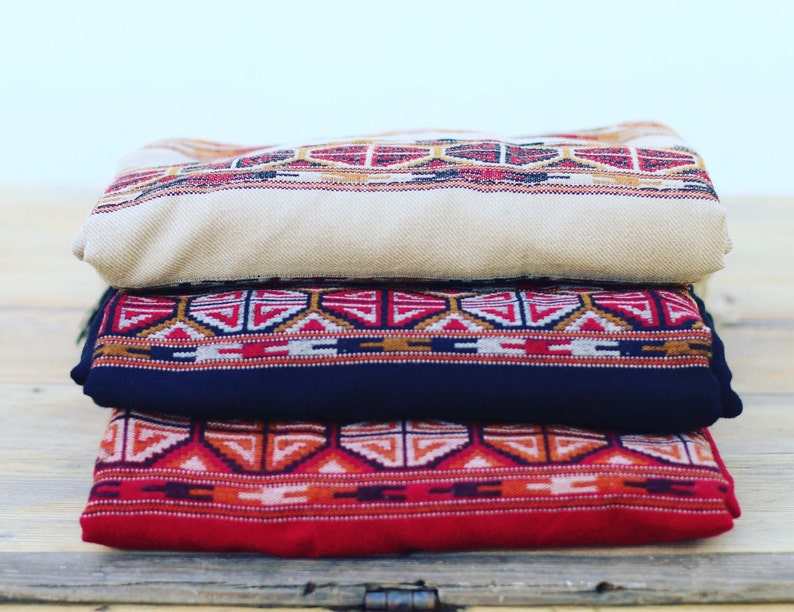 May include: Three woven blankets stacked on top of each other. The top blanket is beige with a red and brown geometric pattern. The middle blanket is dark blue with a red, white, and brown geometric pattern. The bottom blanket is red with a white, orange, and brown geometric pattern.