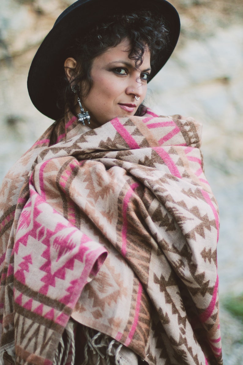 May include: A woman wearing a black hat and a patterned shawl. The shawl features a geometric design in shades of pink, brown, and beige. The shawl is wrapped around the shoulders.