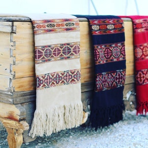 May include: Three woven shawls with fringe, in cream, navy blue, and red, with geometric patterns, draped over a wooden chest.