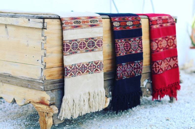 May include: Three woven shawls with fringe, in cream, navy blue, and red, with geometric patterns, draped over a wooden chest.