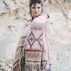 May include: A beige and purple patterned shawl with fringe. The shawl features geometric designs and is draped over the shoulders. The person is wearing blue jeans. The background is a rocky, neutral-toned cliff.