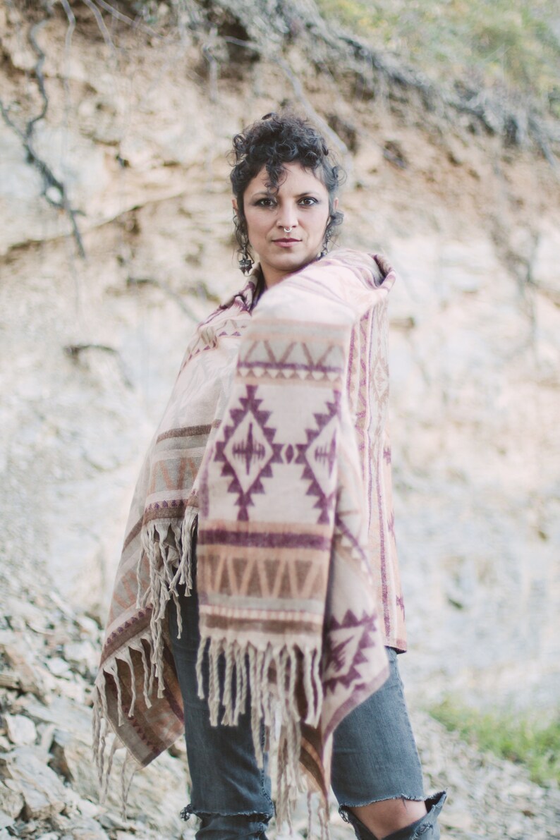 May include: A beige and purple patterned shawl with fringe. The shawl features geometric designs and is draped over the shoulders. The person is wearing blue jeans. The background is a rocky, neutral-toned cliff.