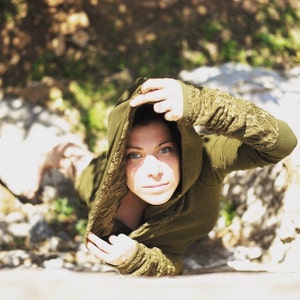 May include: A woman wearing a green hooded sweatshirt with lace detailing on the sleeves. The hood is pulled over her head, and she is looking at the camera.