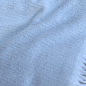 May include: A white, soft, and fluffy throw blanket with a subtle texture. The blanket has a fringe border.