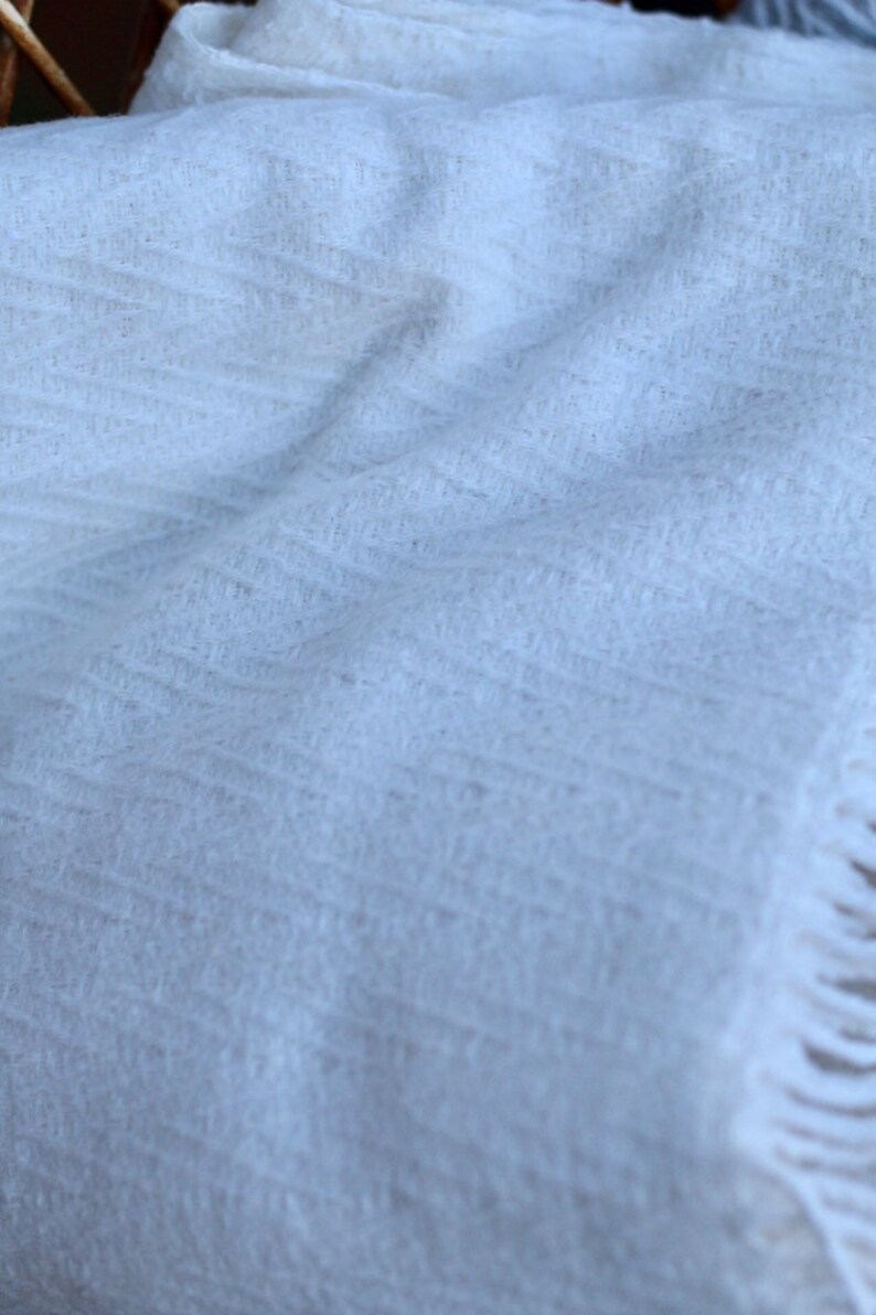 May include: A white, soft, and fluffy throw blanket with a subtle texture. The blanket has a fringe border.