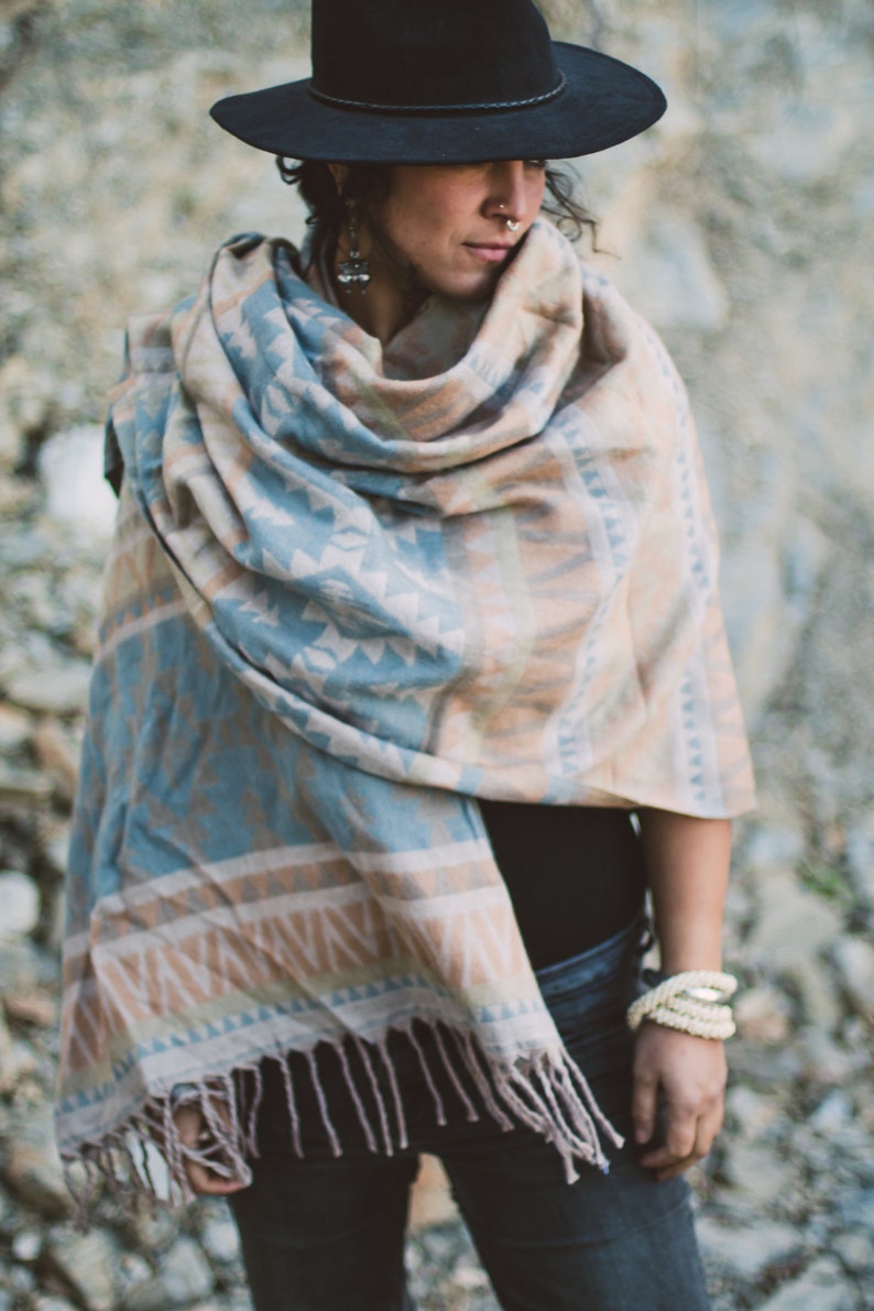 May include: A person wearing a black hat and a fringed shawl. The shawl features a geometric pattern in shades of blue, tan, and white. The person is also wearing a bracelet.