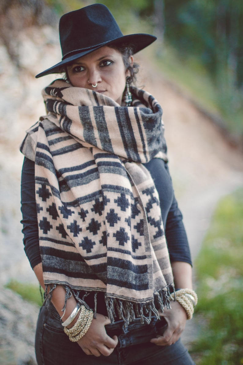 May include: A person wearing a black fedora hat and a beige and black patterned scarf. The scarf has a geometric design and is draped around the neck. The person is also wearing bracelets and a black long-sleeve shirt.