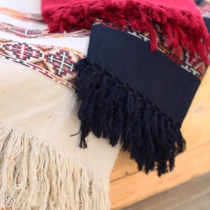 May include: Three woven blankets with fringe. The top blanket is red with white and black geometric patterns. The middle blanket is dark blue with white and red geometric patterns. The bottom blanket is cream with white and red geometric patterns.