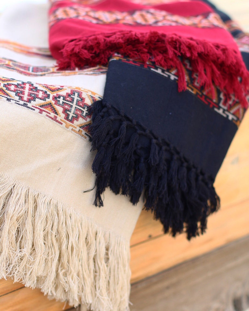 May include: Three woven blankets with fringe. The top blanket is red with white and black geometric patterns. The middle blanket is dark blue with white and red geometric patterns. The bottom blanket is cream with white and red geometric patterns.