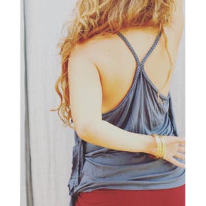 May include: A woman wearing a blue tank top with a criss-cross strap design. She is wearing multiple gold bracelets on her wrist.