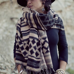 May include: A black fedora hat, a black long-sleeve shirt, and a patterned scarf with black, tan, and gray geometric designs. The scarf is fringed at the ends. Silver and beaded bracelets are worn on the wrists.