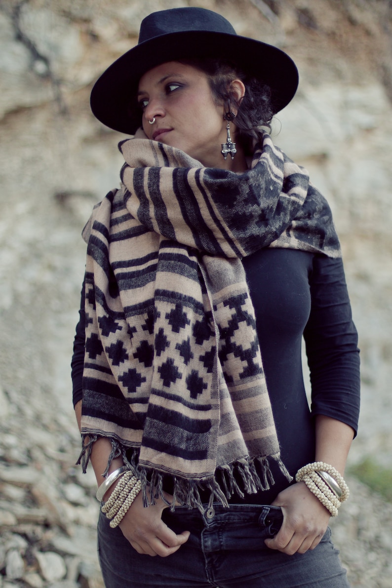 May include: A black fedora hat, a black long-sleeve shirt, and a patterned scarf with black, tan, and gray geometric designs. The scarf is fringed at the ends. Silver and beaded bracelets are worn on the wrists.
