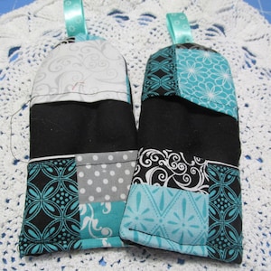 May include: Two quilted oven mitts with a patchwork design. The mitts feature black, gray, and turquoise fabrics with floral and swirl patterns. Each mitt has a black center and a turquoise ribbon loop.