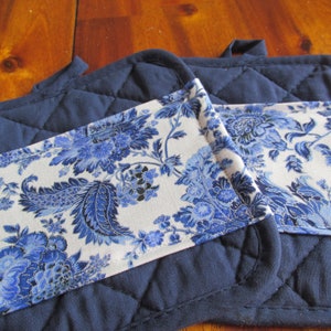 May include: Two blue quilted pot holders with a white floral fabric trim. The floral fabric features a blue and white paisley pattern.